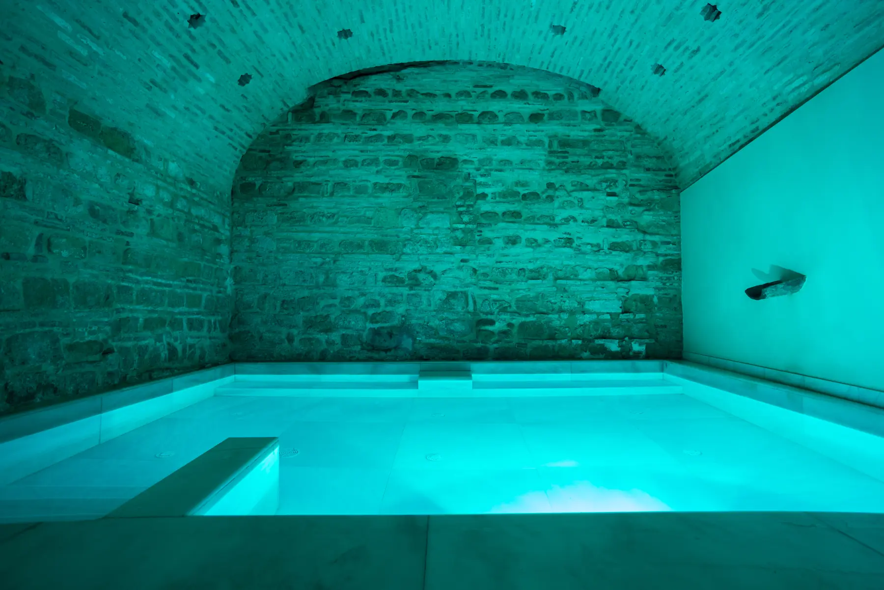 Hammam con bóveda de ladrillo iluminada en turquesa y piscina de mármol blanco en los Baños Árabes de Córdoba
